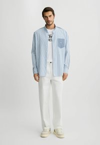 Striped blå og hvid skjorte med en lomme, båret over en hvid grafisk t-shirt, parret med hvide bukser og cremefarvede sneakers.