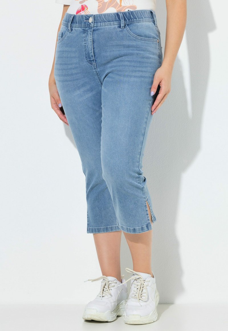 Nainen, jolla on vaaleansiniset lyhennetyt farkut sivuhalkioilla ja valkoiset lenkkarit, seisoo yksiväristä valkoista taustaa vasten.