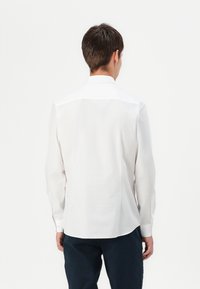 Homme aux cheveux courts et foncés, portant une chemise blanche à manches longues et un pantalon foncé, vu de dos sur un fond uni.