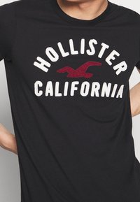 Camiseta de algodón negra con el texto bordado "HOLLISTER CALIFORNIA" y un logo de gaviota color burdeos en el centro. Manga corta, cuello redondo.