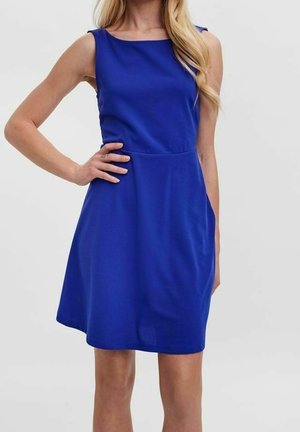 Mouwloze koningsblauwe jurk tot op de knie met een nauwsluitende taille, gedragen door een persoon met één hand op de heup en lang blond haar.