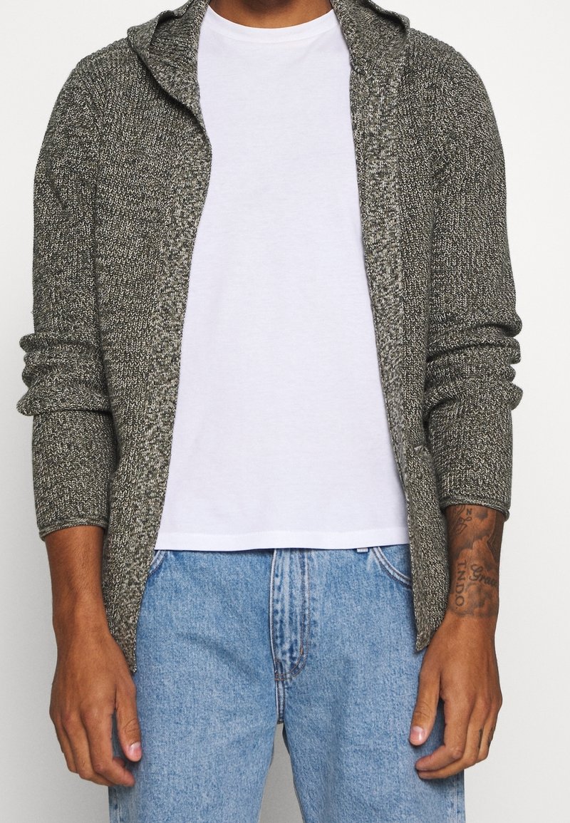 Grauer Strickcardigan mit Kapuze, offenem Vorderteil und langen Ärmeln, getragen über einem weißen T-Shirt, kombiniert mit hellblauen Jeans.