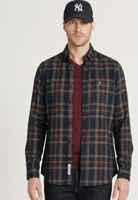 Camisa de cuadros en azul marino y rojo con diseño abotonado, mangas largas y un logo sutil. Se lleva sobre una camiseta de cuello en V color granate.