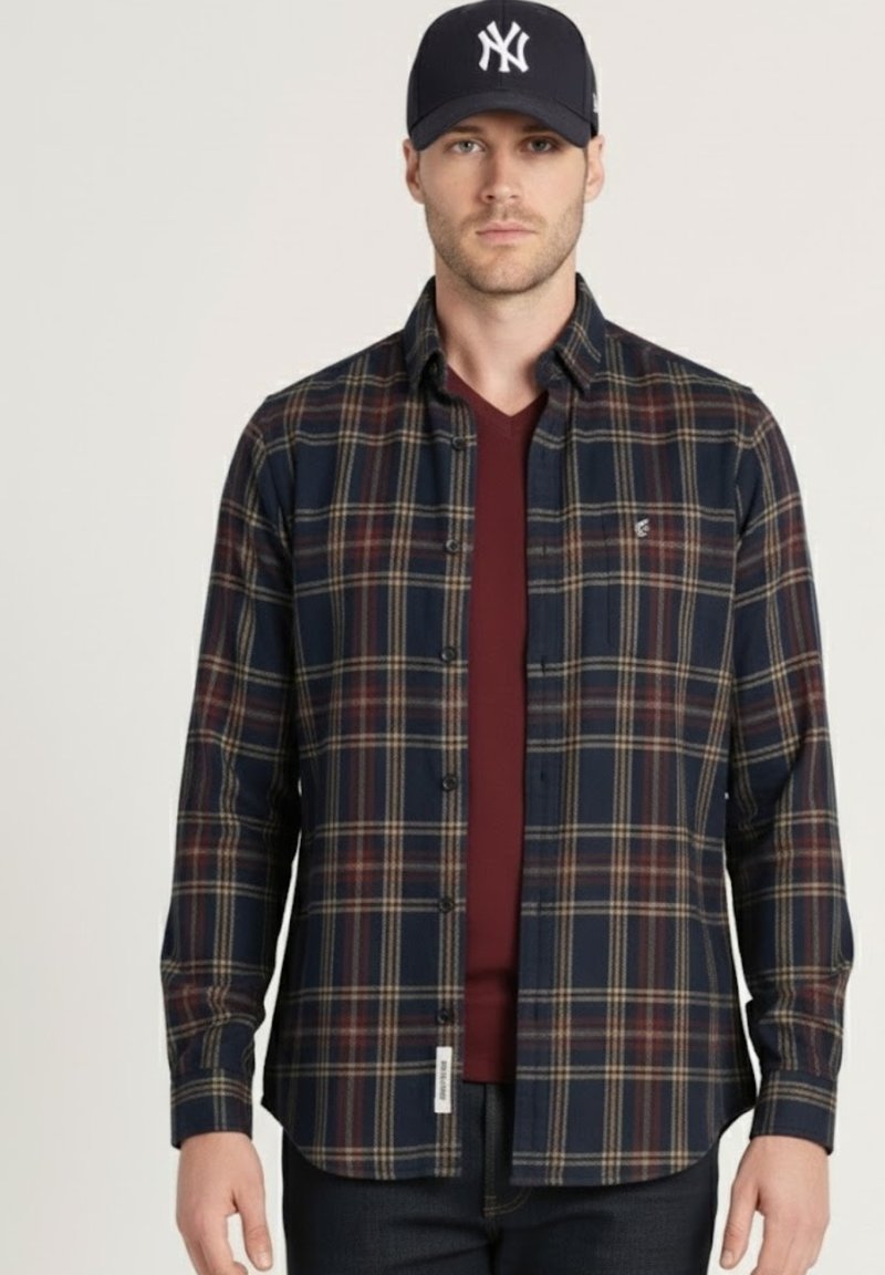 Camisa de cuadros en azul marino y rojo con diseño abotonado, mangas largas y un logo sutil. Se lleva sobre una camiseta de cuello en V color granate.