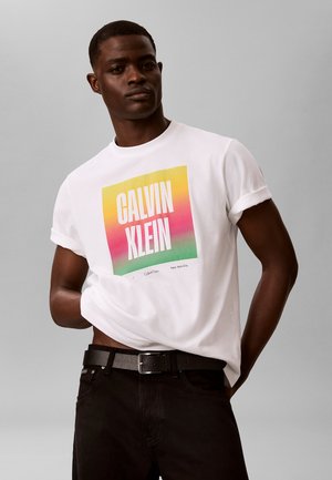 Άνδρας που φοράει λευκό T-shirt με γυρισμένα μανίκια και πολύχρωμο λογότυπο Calvin Klein, μαύρη ζώνη και μαύρο παντελόνι, στέκεται μπροστά σε απλό γκρι φόντο.