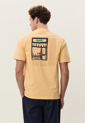 Mann von hinten, trägt ein senfgelbes T-Shirt mit einer Café-Schaufenster- und Textgrafik auf dem Rücken, kombiniert mit dunkler Hose.