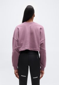 Κοντή μωβ φούτερ με υπερμεγέθη μανίκια, ριμπ πατιλέτα λαιμού και λογότυπο στο πίσω μέρος, σε συνδυασμό με μαύρο κολάν με αντανακλαστικές λεπτομέρειες.