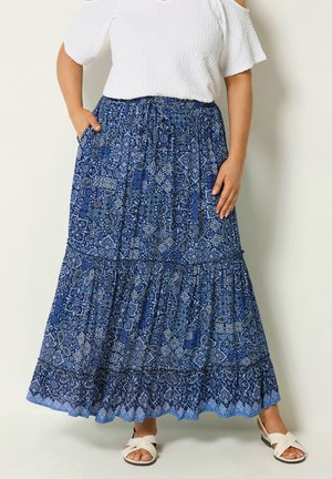 Kvinde iført en blå mønstret lang nederdel, hvid cold-shoulder top og hvide sandaler, stående med den ene hånd i lommen foran ensfarvet baggrund.