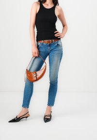 Woman wearing black tank top and blue jeans with brown belt, holding a brown handbag, and wearing black pointed-toe heels.
