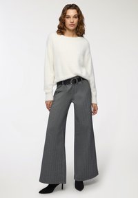 Kobieta ubrana w biały puszysty sweter, szare szerokie spodnie z czarnym paskiem oraz czarne szpilki z ostrymi noskami, stojąca na jednolitym tle.