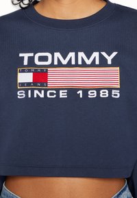 Sudadera recortada en azul marino con el texto bordado en blanco "TOMMY JEANS" y un logotipo rectangular en diseño de franjas rojo, blanco y azul.