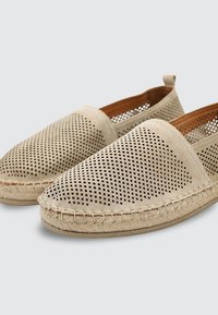 Par de alpargatas beige sin cordones con parte superior perforada y suela trenzada de yute, fotografiadas sobre un fondo gris claro.
