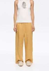 Light-colored tank top with a graphic print, paired with loose-fitting, light yellow linen pants and light beige sneakers.