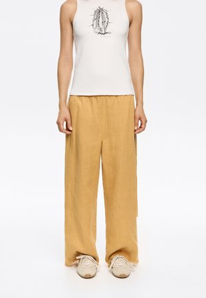 Light-colored tank top with a graphic print, paired with loose-fitting, light yellow linen pants and light beige sneakers.