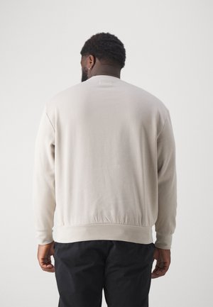 Homme à la peau foncée et aux cheveux courts bouclés, portant une chemise beige à manches longues et un pantalon noir, debout de dos sur un fond blanc.