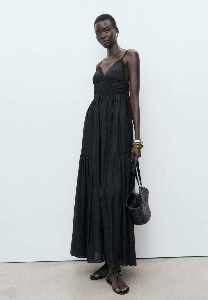 Robe maxi noire avec des bretelles spaghetti et un corsage ajusté. Présente des couches de tissu étagées, une texture tissée, et assortie à un sac noir.