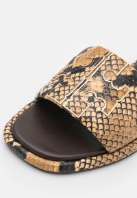 Gros plan d'une sandale mule à motif peau de serpent marron et noir, avec un logo décoratif doré sur la bride et une semelle intérieure noire.