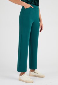 Teal, wijde broek met een gladde textuur en zijzakken. Heeft een hoge tailleband en subtiele zijsplits aan de zoom. Gemaakt met lichte sneakers.