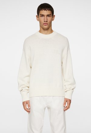 Maglione a maglia color crema con collo rotondo, polsini e orlo a costine. Presenta un logo discreto sul petto. Texture liscia, vestibilità rilassata.