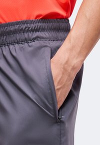 Main reposant dans la poche latérale d’un short de sport gris à taille élastique porté avec un t-shirt rouge.