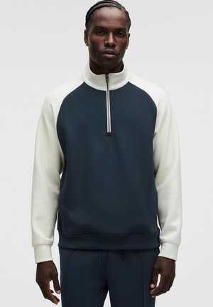 Hombre con cabello trenzado que lleva un jersey de manga larga azul marino y blanco con cuello alto y cremallera frontal, de pie frente a un fondo liso.
