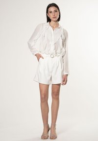 Blusa bianca con volant a maniche lunghe, abbinata a pantaloni corti bianchi su misura con una cintura dotata di una fibbia circolare argentata e sandali con tacco basso.