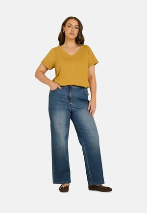 Kvinde står med den ene hånd i lommen iført en sennep kortærmet V-hals top, blå vidbenede jeans og brune balletsko.
