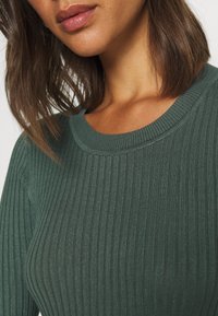 Maglione a coste verde scuro con scollatura tonda e design aderente. Il tessuto testurizzato e la leggera lucentezza ne accentuano l'aspetto.