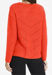 Rød strikket sweater med en chevronmønster og åbne detaljer, rund halsudskæring og dropped skuldre, parret med sorte bukser.