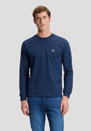 Ung mand iført en navyblå langærmet skjorte med et lille "lee" logo og blå jeans står foran en ensartet lys baggrund.