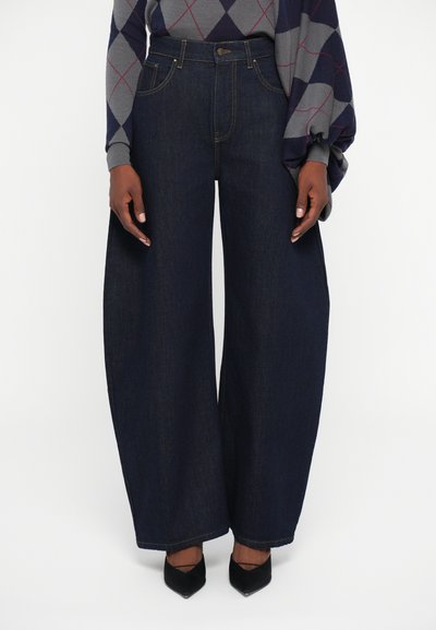 Person wearing dark blue wide-leg jeans, a gray and navy argyle sweater, and black pointed-toe heels standing against a white background.