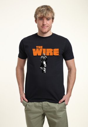 Czarna koszulka z napisem "THE WIRE" w pomarańczowym kolorze, z czarno-białą grafiką mężczyzny w płaszczu trench. Materiał bawełniany, krótkie rękawy.