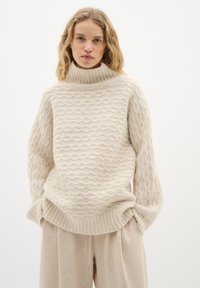 Flauschiger, cremefarbener Strickpullover mit hohem, geripptem Kragen, der ein wellenförmiges Texturmuster aufweist, kombiniert mit lockeren, beigen Hosen.
