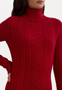 Pulover roșu tricotat cu gât înalt, având un model cu cabluri, accente ribbate și mâneci ajustate. Textura este groasă și călduroasă.