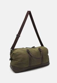Bolsa de lona verde con detalles en cuero marrón, cierre superior con cremallera, asas dobles y correa de hombro ajustable. Textura suave, forma redondeada.