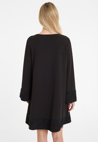 Schwarzes Kleid mit langen Ärmeln, lockerem Schnitt und texturierten Fransenakzenten an den Manschetten und dem Saum, die einen geschichteten visuellen Effekt erzeugen.
