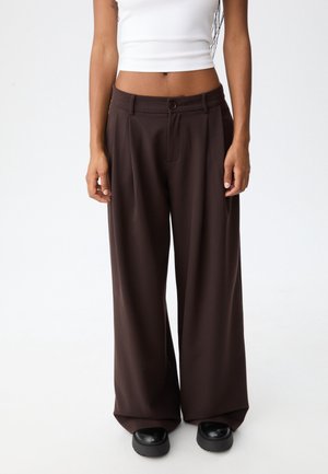 Vrouw met losse donkerbruine wijde broek, wit cropped topje en zwarte platformschoenen, staand tegen een effen achtergrond.