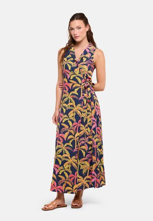 Mujer con vestido cruzado sin mangas de color azul marino con estampado de palmeras en amarillo, rosa y naranja y sandalias planas marrones, de pie frente a un fondo liso.