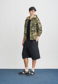 Kameru jaka ar kapuci, melna T-kreklu, melnas šortus un sporta apavus, uz neitrāla fona. Tekstūrēts un sportisks dizains.