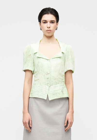 Blouse vert clair texturée avec des manches courtes bouffantes, un col carré et une fermeture à boutons à l'avant. Portée avec une jupe grise.