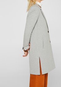 Persona rubia con abrigo gris hasta la rodilla con abertura, suéter blanco de cuello alto y pantalones naranjas, de perfil contra un fondo liso.