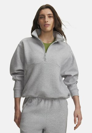 Vrouw met lang bruin haar die een lichtgrijze halve rits trui draagt en bijpassende trainingsbroek met elastische tailleband en zakken.