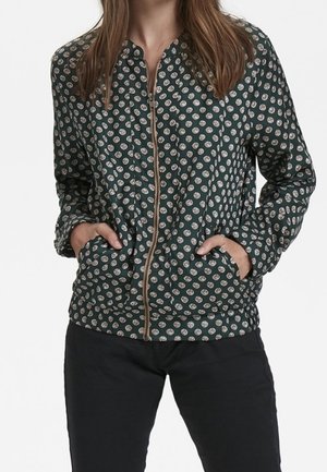 Žena oblečená v tmavě zelené bundě na zip s bílými kruhovými vzory a černými kalhotami, ruce zasunuté v kapsách bundy.