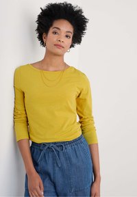 Kollane pikade varrukatega särk ümmarguse kaelusega, millele on sobitatud sinised paeltega püksid suurte taskutega. Sile tekstuur, juhuslik lõige.