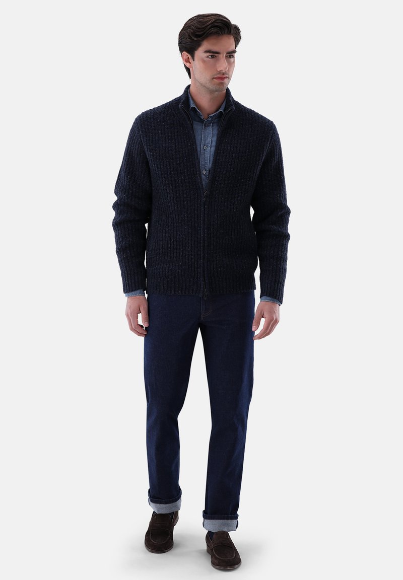 Marineblauer Cardigan mit geripptem Textil, Reißverschluss und Schalkragen, getragen über einem Jeanshemd und kombiniert mit hochgekrempelten dunklen Jeans und braunen Schuhen.