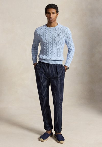 Άνδρας που φοράει ανοιχτό μπλε πουλόβερ πλεκτό με σχέδιο, μπλε παντελόνι με λεπτές ρίγες και μπλε παπούτσια slip-on, όρθιος με τα χέρια στις τσέπες.