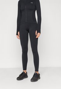 Μαύρο εφαρμοστό αθλητικό μπουφάν με μακριά μανίκια, κλείσιμο με φερμουάρ και θήκες για τον αντίχειρα, σε συνδυασμό με μαύρες εφαρμοστές κολάν και μαύρα αθλητικά παπούτσια.