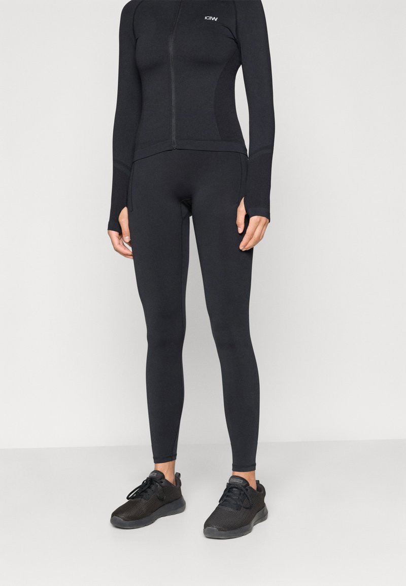 Μαύρο εφαρμοστό αθλητικό μπουφάν με μακριά μανίκια, κλείσιμο με φερμουάρ και θήκες για τον αντίχειρα, σε συνδυασμό με μαύρες εφαρμοστές κολάν και μαύρα αθλητικά παπούτσια.