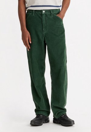 Osoba ubrana w ciemnozielone sztruksowe spodnie i czarne trampki, z jedną ręką w kieszeni i częściowo widoczną koszulą w paski.