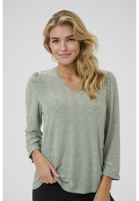 Haut à manches longues vert clair avec un décolleté en V, doté d'un tissu texturé et d'un motif à œillets. Design doux et fluide avec un ourlet arrondi.